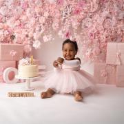 Baby beim Cake-Smash-Fotoshooting zum ersten Geburtstag, sitzend vor einer rosa Blumenwand mit Torte, Geschenkboxen und dem Namen „Kehlani“.