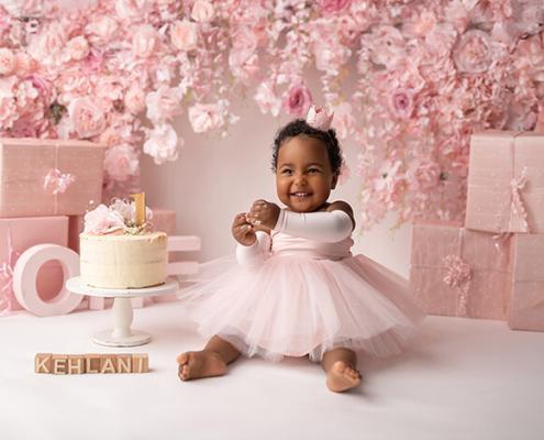 Baby beim Cake-Smash-Fotoshooting zum ersten Geburtstag, sitzend vor einer rosa Blumenwand mit Torte, Geschenkboxen und dem Namen „Kehlani“.