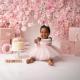 Baby beim Cake-Smash-Fotoshooting zum ersten Geburtstag, sitzend vor einer rosa Blumenwand mit Torte, Geschenkboxen und dem Namen „Kehlani“.