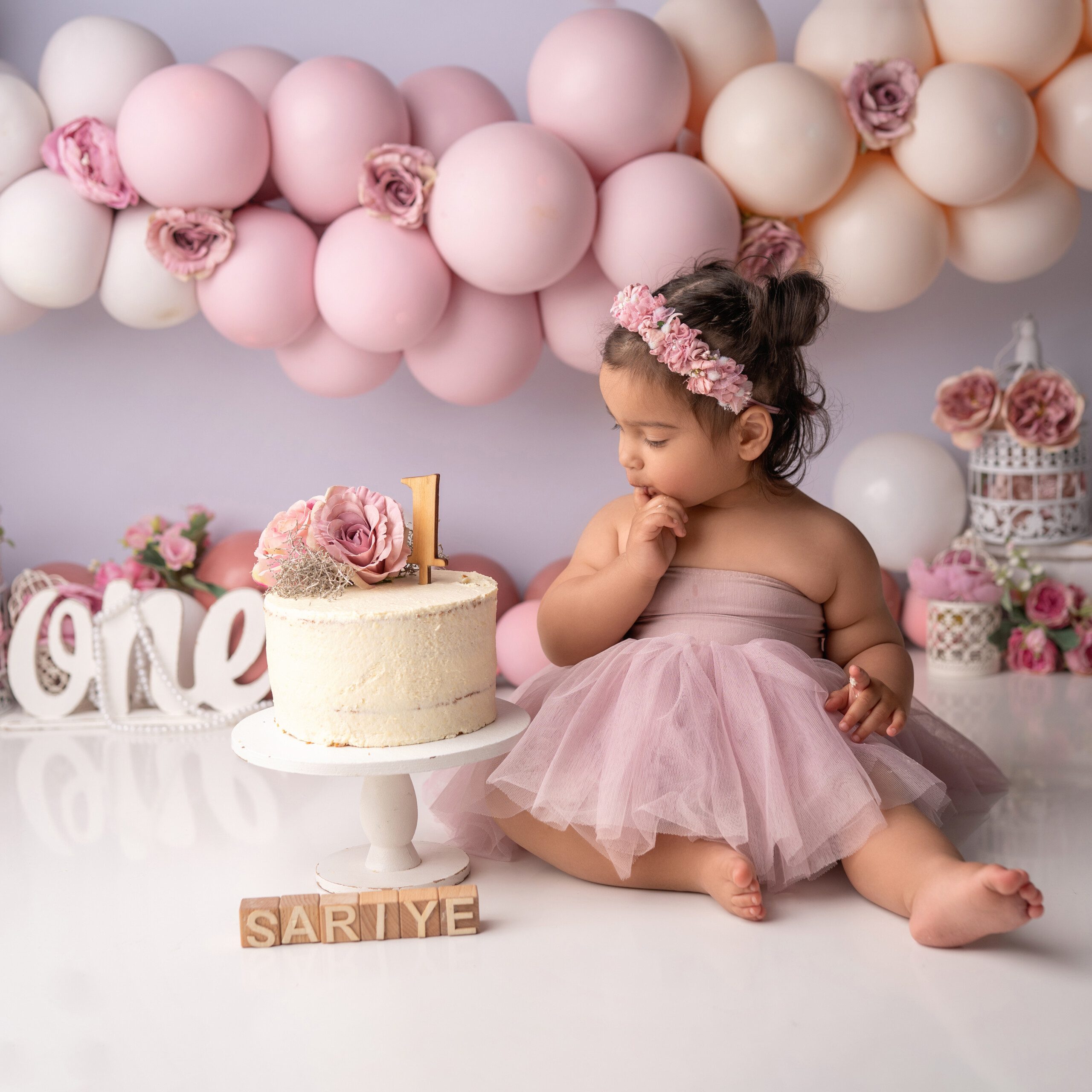Baby beim Cake-Smash-Fotoshooting zum ersten Geburtstag, umgeben von rosa Ballons und Blumen, mit einer personalisierten „ONE“-Dekoration und dem Namen „Sariye“.